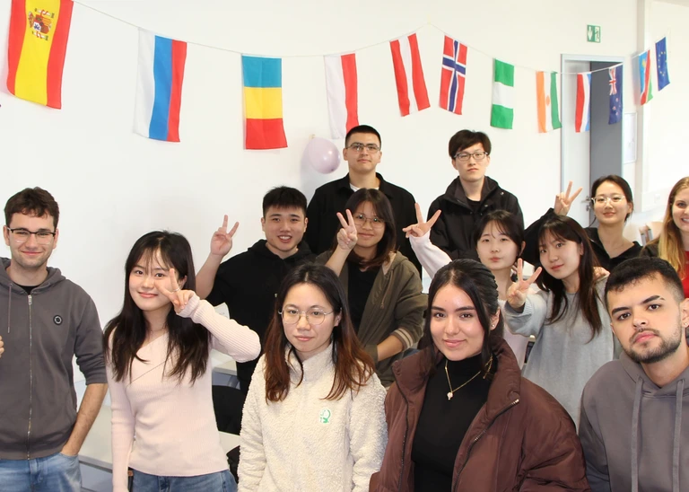 A group of 13 young people smiling into the camera, with various national flags decorated on the wall in the background. 